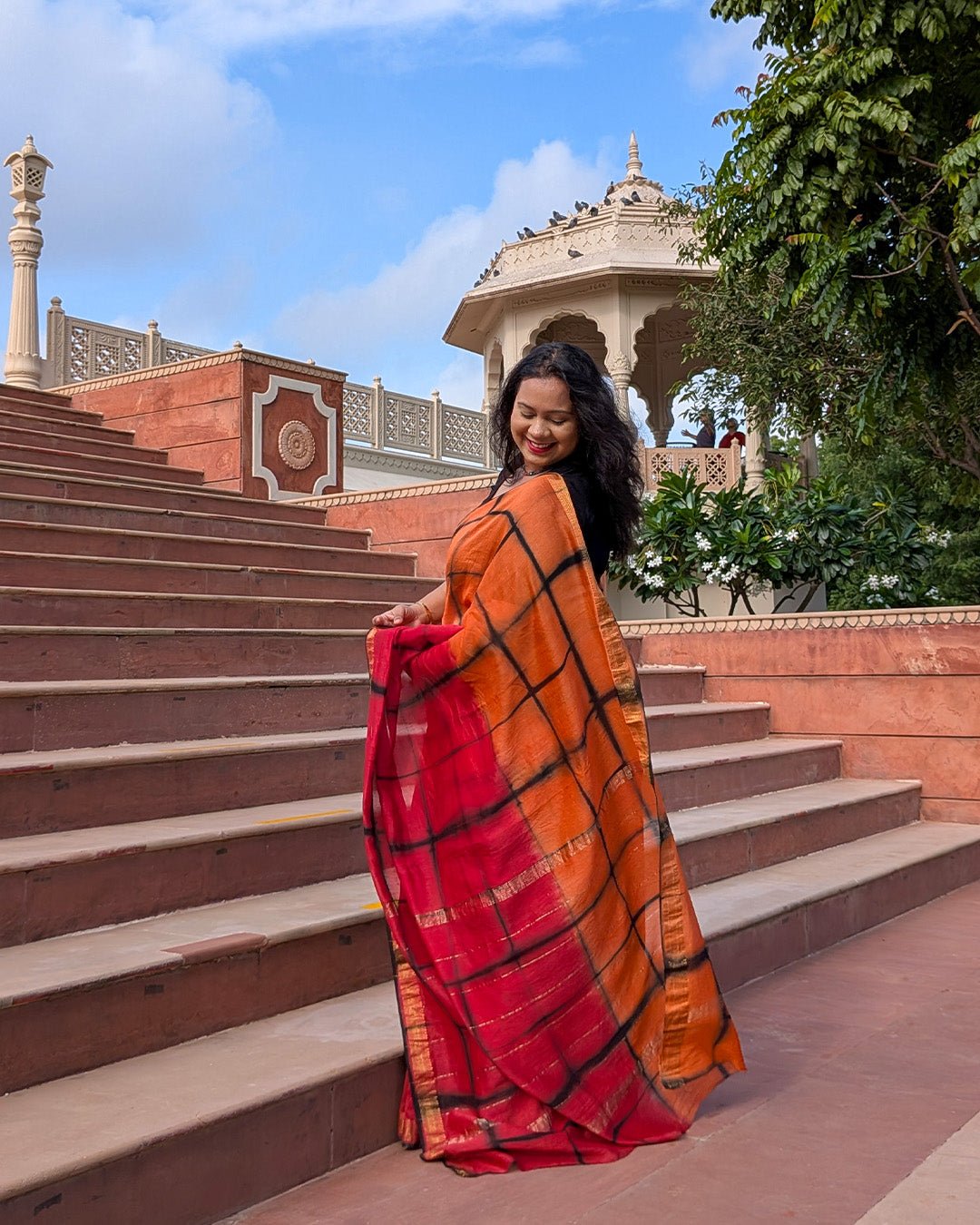 Model posing on outdoor stairs wearing the orange and red checked Maheshwari handwoven saree by House of Ojasvi.