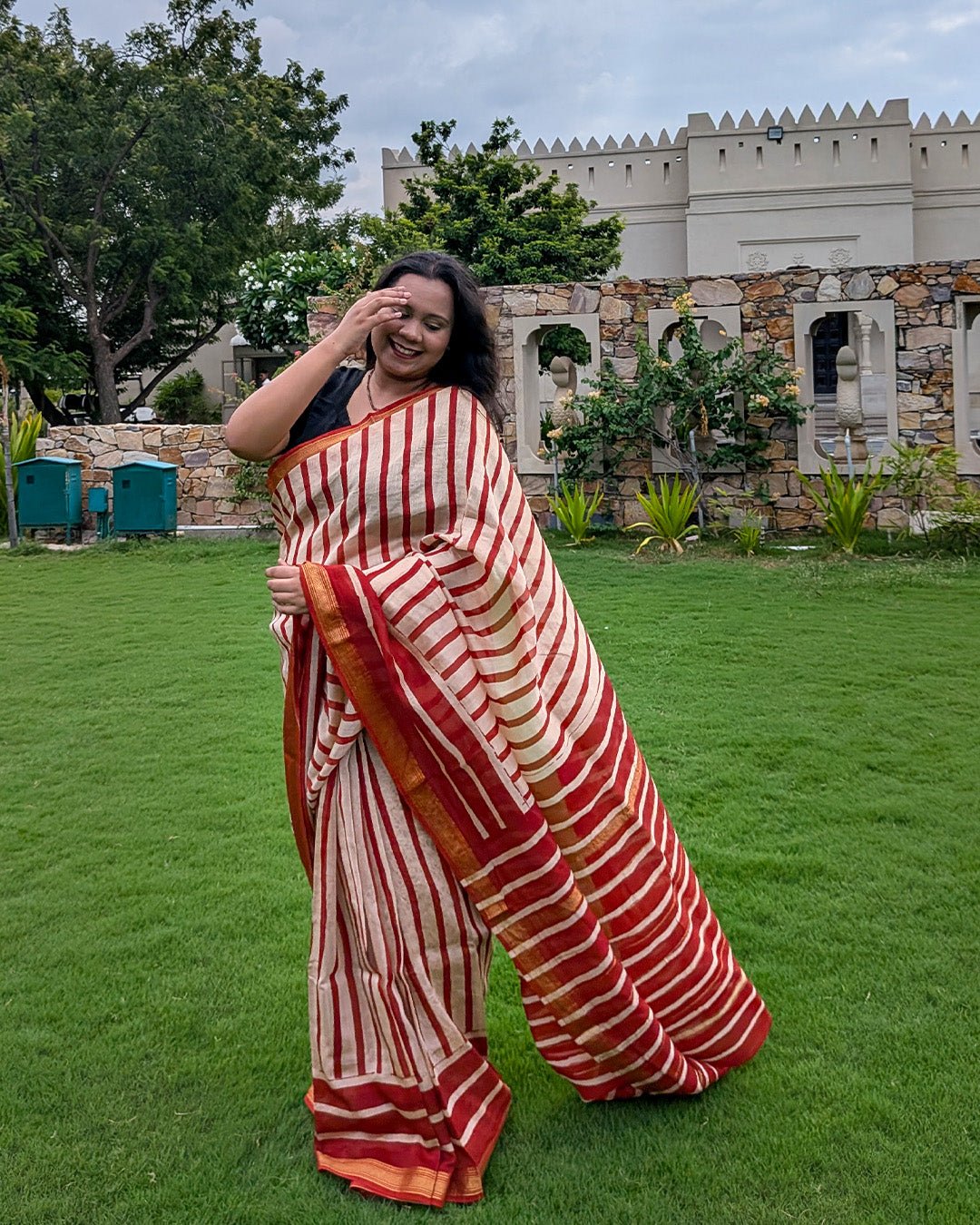 Model posing gracefully against a wooden doorway wearing the red and cream striped Maheshwari handwoven saree by House of Ojasvi.