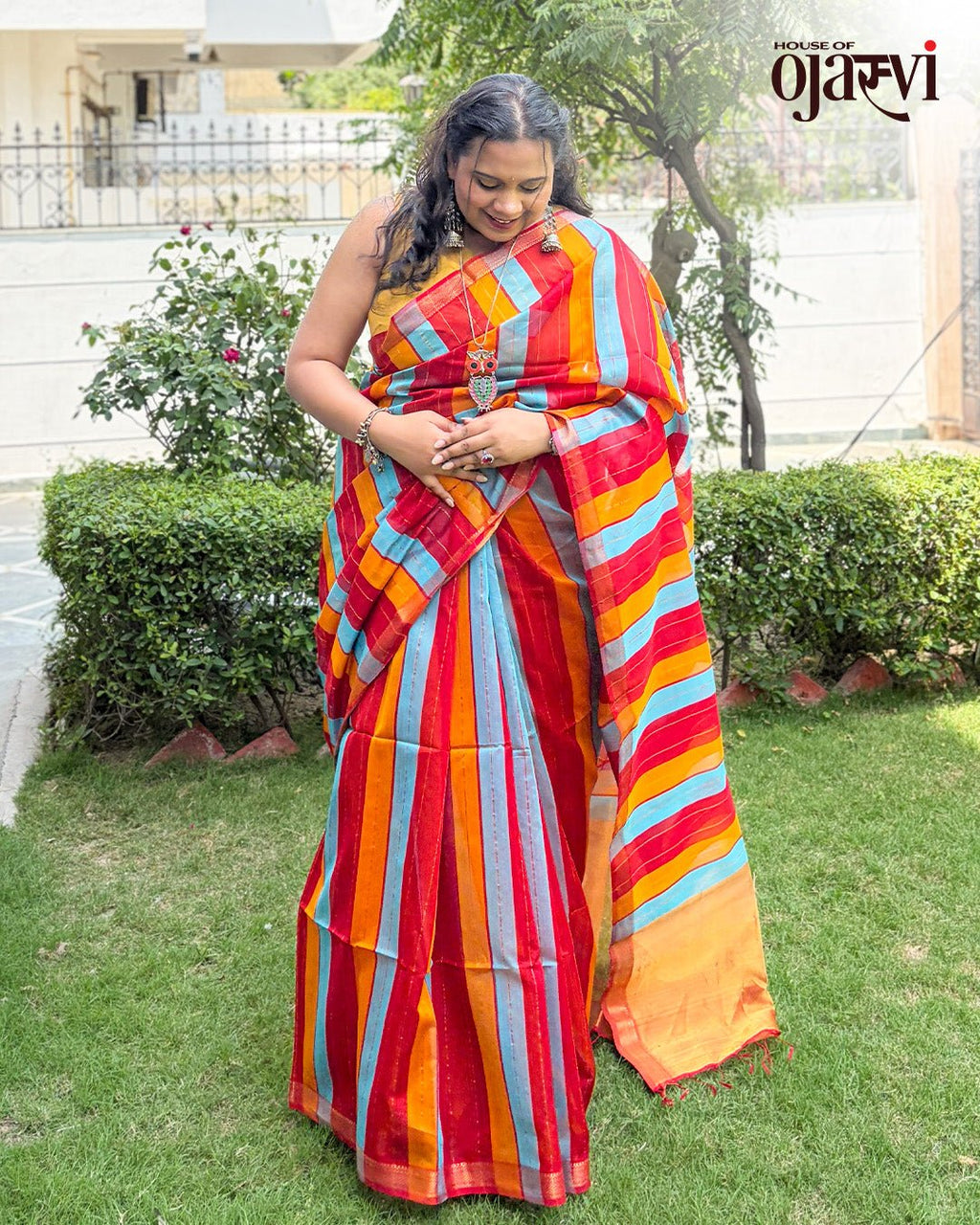 Model posing outdoors showcasing the rich gold zari border of the red, blue, and orange striped Maheshwari saree by House of Ojasvi.