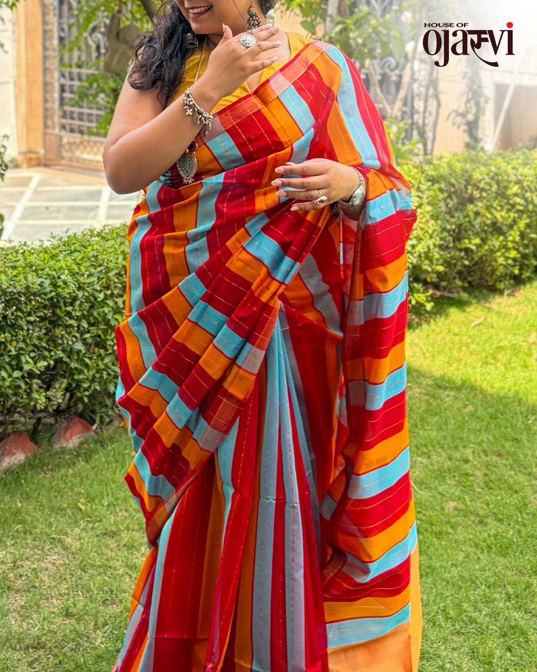 Outdoor garden view of a model styling the red, blue, and orange striped Maheshwari handwoven saree.